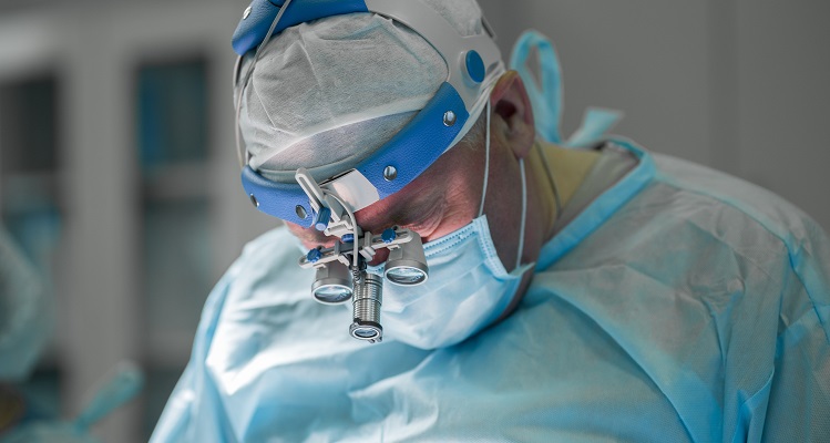 Surgeon performing plastic surgery on breasts in hospital operating room. Surgeon in mask wearing loupes during medical procedure. Breast augmentation, enlargement, enhancement